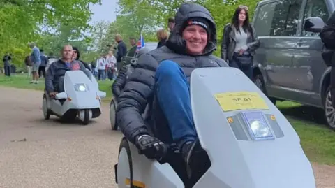 Bryan Griffin Bryan Griffin is smiling as he rides in his C5 vehicle. He is wearing a padded anorak, blue trousers and gloves. There are other C5s behind him and a woman is walking past some parked cars next to the pathway