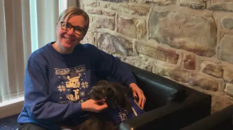 A woman with short blonde hair and glasses, who is wearing a blue Christmas jumper with a dog on it, is holding a black dog while sitting on a lounge chair. 