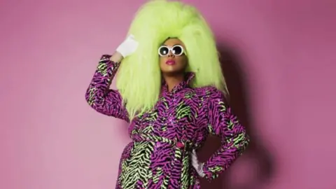 A black man with sunglasses in a large yellow wig and patterned pink and yellow dress against a pink background