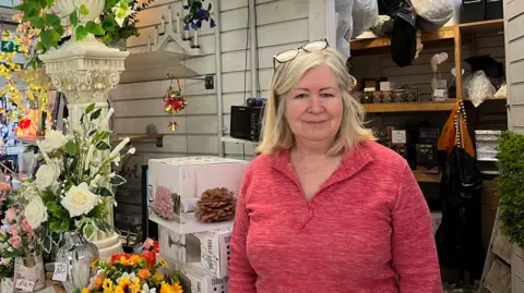 Jonny Humphries/BBC Lisa Croston, 64, who has blonde shoulder length hair, smiles at the camera in front of a stall filled with floral arrangements and ornaments. She is wearing a pair of glasses resting on the top of her head, and a pink quarter-zip jumper. 