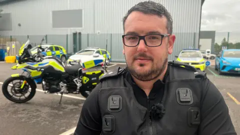 Matt wearing his police uniform and black rimmed glasses, with short dark hair smiling at the camera. He is standing in a police car park with police cars and a police motorbike in the background.