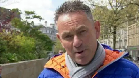 BBC Marcus Farmer has brown hair with grey temples and wears a blue anorak with orange lining and a grey scarf.