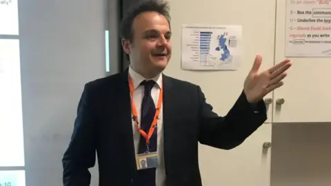 Tristan Osborne Tris Osbourne is wearing a suit with an orange lanyard around his neck, giving a talk in an office.