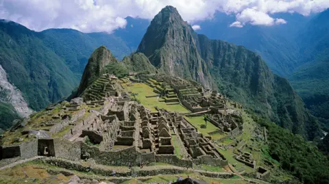 Machu Picchu is a small town consisting of stone houses and walls that have been worn down over time. It is built on a steep mountain ridge between green peaks.