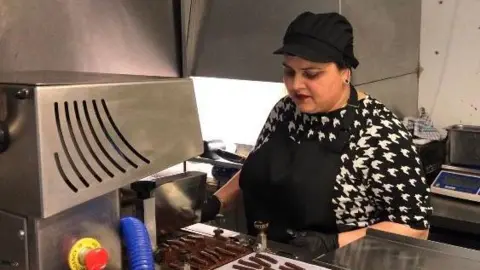 A woman standing behind a silver machine in a kitchen. There are thin chocolates in front of her on the metal work surface. The woman is wearing a black hat and apron as well as black gloves. She is looking down at the chocolates. 