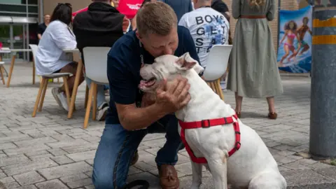 Utilita Tilly is sat looking away from the camera wearing her red harness. Shaun Dollamore is crouched next to her, he has his hands around her face affectionately and is leaning in to kiss the top of her head. Shaun has short dark blonde hair and wears a navy blue polo shirt with jeans and tan brown leather deck shows. In the background there are people sat around a table talking.
