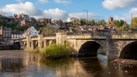 A bridge across a river with a town in the background, including houses, a church and trees.