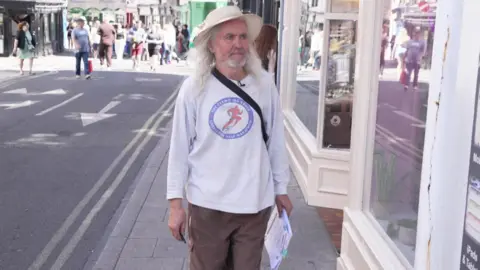 A man wearing a hat and a long sleeved t-shirt walks down a shopping street, holding pieces of paper.