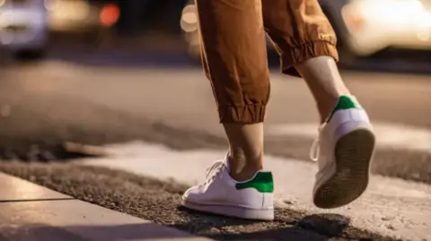 A close up of a young woman's feet as she walks along a road at night. She is wearing white trainers and elasticated light brown leggings which come down to just above her ankles. She is not wearing socks. The background, which shows lights and a car, is out of focus.
