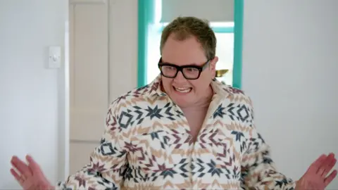 Alan Carr smiles with his hands outstretched. He wears black rimmed glasses and a patterned shirt