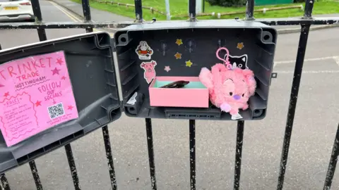 The box is attached to a fence at Bridgford park. There is a pink fluffy animal inside and some stickers. 