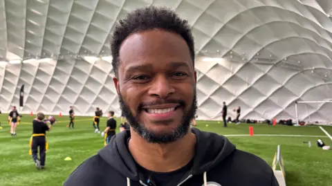 A man with short black hair and facial hair smiling at the camera as he stands in front of kids playing sports. He is wearing a black jacket.