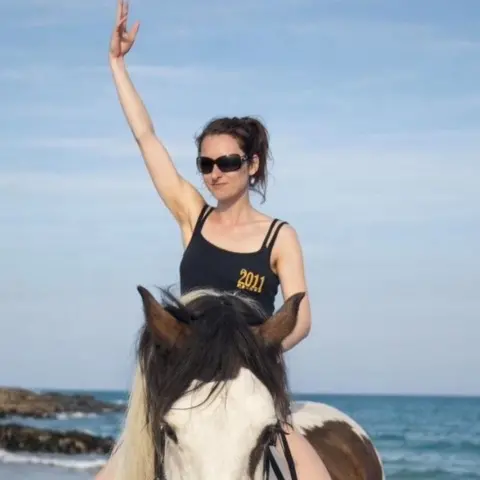Naomi MacIvor family Naomi MacIvor is sitting on a brown and white horse on a beach. Naomi is wearing a dark coloured vest top, sunglasses and has her hair tied up. She has an arm raised.