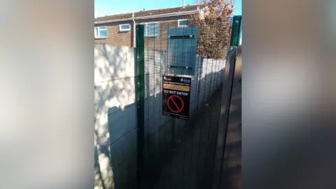 A green metal gate at the start of an alleyway with a "Do not enter sign" on the front.