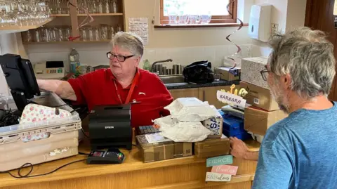 A woman serving a Post Office customer from a counter set up in a village hall. There is a till on the counter, with the produce in bags, and thee are glasses and a sink behind the woman along the back wall.