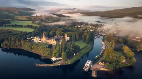 An aerial view of Fort Augustus and Caledonian Canal on the shores of Loch Ness. Mist covers parts of the villages and nearby hills.