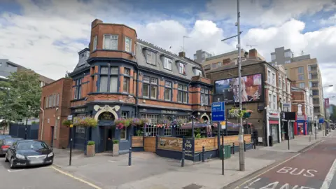 Google Street view of the outside of The Old Hat pub in Ealing, a brick building with a turret, dormer roof and hangings baskets of flowers, with a dark painted frontage and window frames, and a fenced off outdoor area 