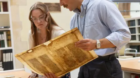 A man and woman hold up an ancient sheet of yellowed text with holes and stains around its edges.