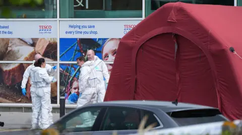 Four forensic workers dressed in white zip-up suits and wearing blue gloves stand outside a red tent outside Fairgreen Shopping Centre in Carlow. Behind the tent are large windows with Tesco signs.