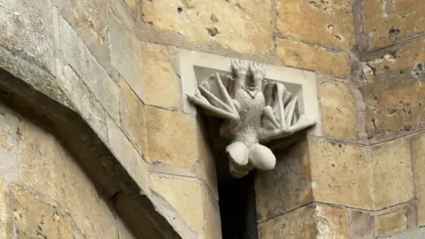 Eleanor Maslin/BBC A stone bat carving is peeking through a dark slit window of a cathedral. Its feet can be seen resting on top of the window with its wings outstretched beside it. The window is cut in a yellow brick wall.
