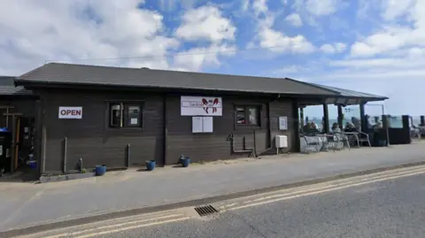 A small seaside café with dark wooden cladding and a sign reading “Sandside Café” sits beside a coastal road. 