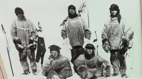 A black and white photo which has three men standing and two more sitting in front of them on the snow. They are wearing heavy clothing and hats and have ski poles. There is a Union Jack flag in the snow behind them.