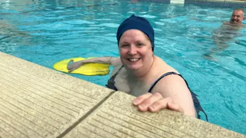 Claire is in a swimming pool. Wearing a navy swimming cap and holding a yellow floater.