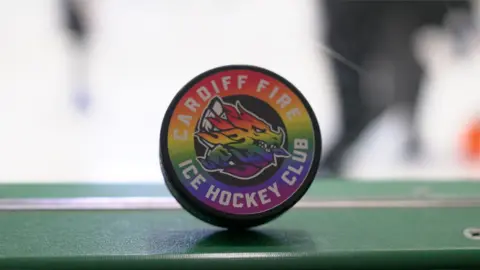 A hockey puck with the multi-coloured rainbow flag spread across it. There is also a dragon emblem on the puck.