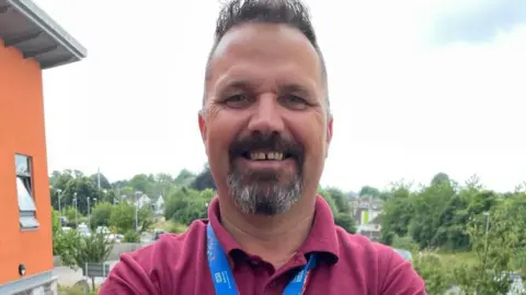 Somerset NHS Foundation Trust Dairin Keating is standing on a balcony wearing a red shirt. He is smiling at the camera. Behind him are trees and houses in the distance.