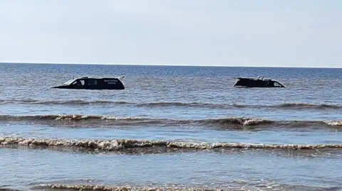 Two trucks are pictured in the sea, and small waves crashing into the shore.