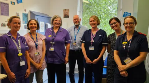 Torbay Hospital A photo which shows seven members of staff standing in a row smiling. The woman to the far left of the photo has a purple hospital uniform on, the woman next to her has a pink shirt on, the woman to the right of her has another purple uniform on. Next to her is a man wearing a blue shirt, and next to him is a woman wearing a blue hospital uniform. The woman on the right of her is wearing a spotty dress and the woman on the far right is wearing a blue hospital uniform. 