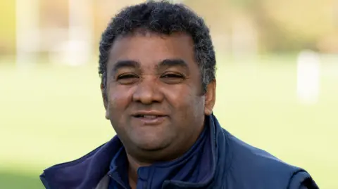 Bedford School Gary Steer, looking at the camera and smiling, wearing a blue top, with a blue gilet. He has short dark hair. 