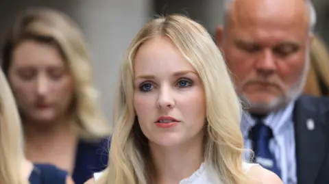 PA Media Lissie Harper speaks at a press conference, with other people in the background. She has light blonde, straight, long hair and wears a white sleeveless top.