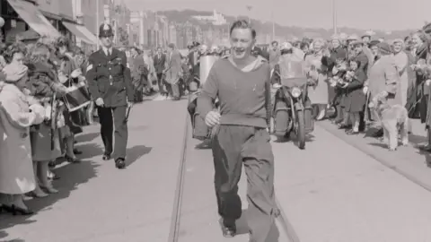 Manx National Heritage A black and white photo of a smiling man walking along some tram tracks. Crowds are gathered around to watch him finish the walking race. A policeman walks behind him and he is followed by a motorbike. 