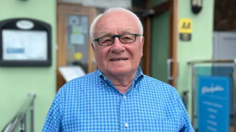 Renzo Martin is an older man wearing a blue, checked shirt and glasses. He is smiling into the camera and standing in front of the Dolphin Hotel.
