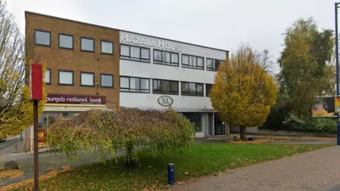 Google A building that is brown on the left and grey on the right, with the words Hazara House on the outside on the highest storey. Trees are in front and to the side of the building. There are some office spaces on the lower storey.