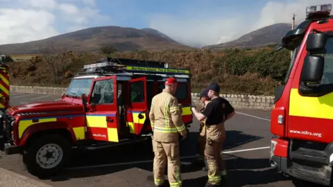The Mourne Mountains can be seen in the distance with smoke from wildfires filling the blue sky. In the foreground three firefighters wearing high vis protective clothing talk. They are standing beside three fire service vehicles. They are red and yellow and one says: "Northern Ireland Fire and Rescue Service".