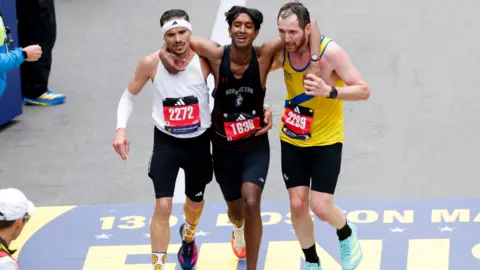 Three men wearing black shorts and different coloured running vests jog towards the finish line of the Boston Marathon with their arms around each other. Robson De Oliveira, who has short, dark hair and a moustasch and is wearing a white vest and a white headband. Ajay Haridasse has short, dark hair and a black vest. Aaron Beggs has short brown hair and a beard and is wearing a yellow vest with a blue stripe.