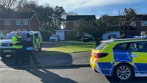 BBC A police car and police van outside a house with white and blue police tape in front of the driveway. There is a car in the driveway and a tree in a small garden