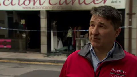 Action Cancer CEO Gareth Kirk standing to the right of the frame, with the damaged Action Cancer store in the background