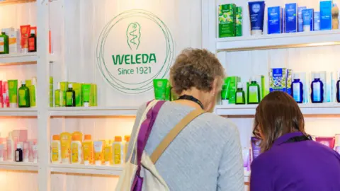 Alamy Two people stand in front of a display shelf featuring various Weleda products arranged by colour: green, yellow, pink, and blue. A circular logo in the centre reads "WELEDA Since 1921" with an abstract plant design above it. The person on the left wears a light grey sweater and carries a tote bag with purple straps; the person on the right wears a purple shirt. They are both facing the shelves so only their backs are visible.