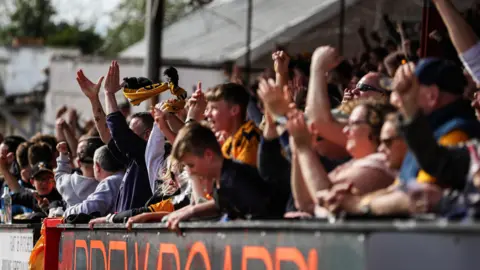 Cambridge United Men, women and children attending a Cambridge United match, in the stands, some cheering and clapping, some wearing the yellow and black club colours