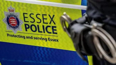 A green and blue Essex Police car with the hip of a police officer in front of it. That officer has a set of handcuffs on them.