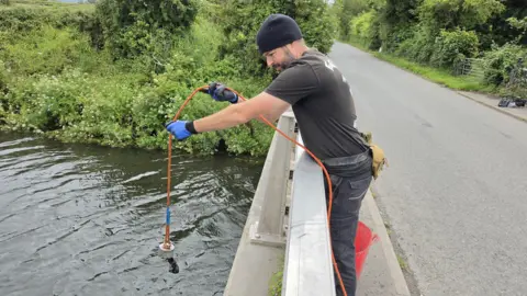 A man stands overlooking a bridge with a river underneath holding an orange rope with a magnet attached which has found some old metal