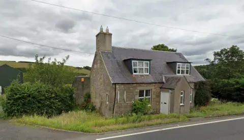 A stone cottage next to a road. The sky is grey and the house is surrounded by a lot of overgrown shrubs and grass