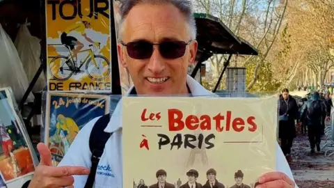 Dino Vescera A man looking the camera wearing sunglasses. He is looking at the camera and smiling. He pointing a poster which says "Les Beatles a Paris". Behind him are other French posters and a street.