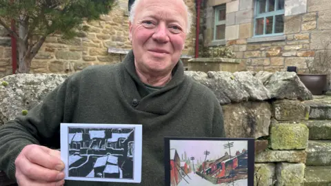HighlightsPR Neville Atkinson, who has short grey hair and is wearing a dark green jumper, is holding up two small pieces of artwork. One, on the left, is a black and white depiction of some back alleys. On the right, red brick houses are depicted under a layer of snow. Mr Atkinson is slightly smiling at the camera as he stands in front of a cottage with a stone wall.