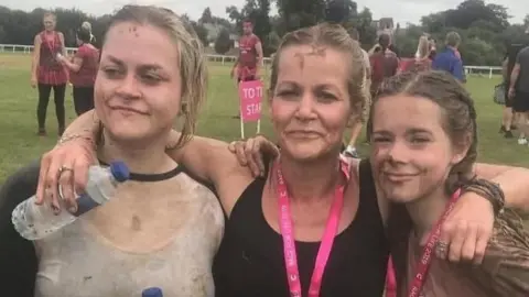 Three women pose for a photo having done a muddy run. The mother is in the middle wearing a black top and medal with a pink lanyard. She has her arms around her two daughters. The one on the right is a teenager, with brown plaited hair and wearing a brown top and a medal. On the left is an older woman in her twenties, wearing a light top and holding a water bottle.