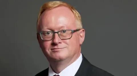 A close-up picture of Luke Akehurst. He has short red hair and is wearing dark-rimmed glasses. The Houses of Parliament can be seen out of focus behind him.