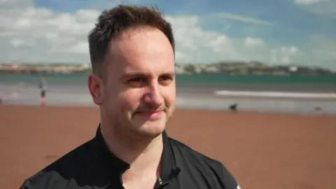 Rev Matt Bray is smiling and looking to the right. Behind him is Paignton beach.
